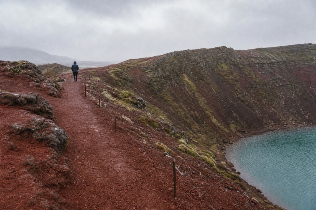 Red volcanic slopes and aquamarine lake of Kerið crater seen from the rim path in drizzle